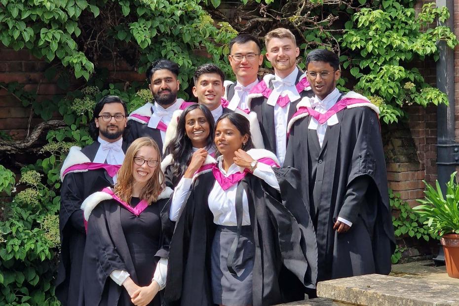 Girton medics students graduating in gowns, pink satin and white fluffy hood. Stood in front of green foliage.