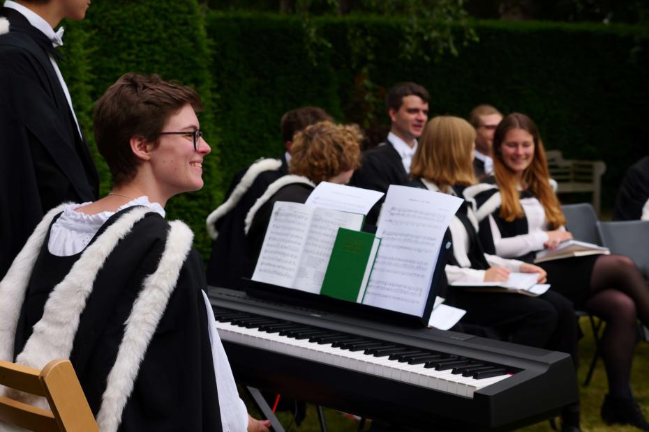 student pianist sitting outside with a keyboard in a graduation gown in the foreground. In the background there are Choir members sitting in conversation with music sheets on their laps, also in graduation gowns