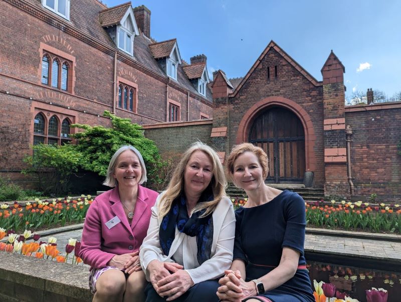Dr Fiona Cooke, Pauline Acerini, and Dr Elisabeth Kendall sit on a low wall in the gardens of Girton College