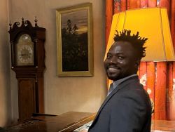 Dr Stephen Oppong Peprah pictured in the Fellows' Drawing Room. A piano, grandfather clock, artwork and a lamp are behind him.