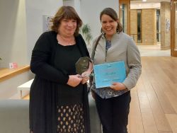 Maureen Hackett stands next to Dr Claire White in a reception space. Maureen holds a glass trophy and Claire holds a certificate.