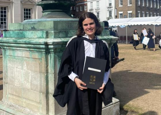 Sophie, a young woman, is smiling and holding a degree certificate outside of Senate House, Cambridge
