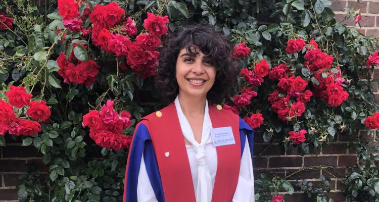 Soudabeh Imanikia in her academic gown (red and blue) in front of a green leafed plant covered with red clustered flowers