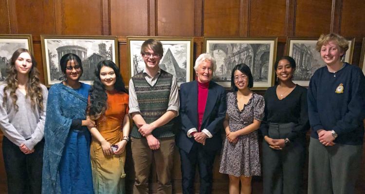 all 7 participants of the 2024 Mountford Prize, stood in a line, with Dr Mountford in the centre. Wood panelling background where 5 black and white scenic landscape pictures hand in a gold frame