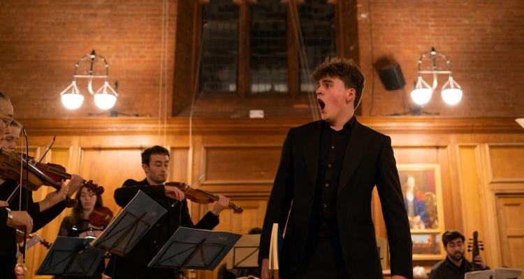 male in the forefront singing, in the background there is string musician's playing in a standing and seated positions. All musicians are dressed in black attire. Room is light with soft yellow lighting creating a ambiance