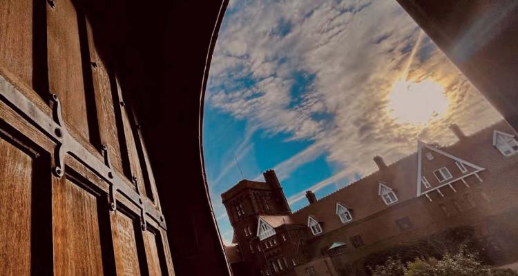 Cropped cover art for the publication 'The Year 2022-23' (College wooden gate door opens out onto courtyard with the view of the College Tower in the glinsing sunshine). Photograph called 'The afternoon of hope' by Ting Zhou (2021 MPhil Education)